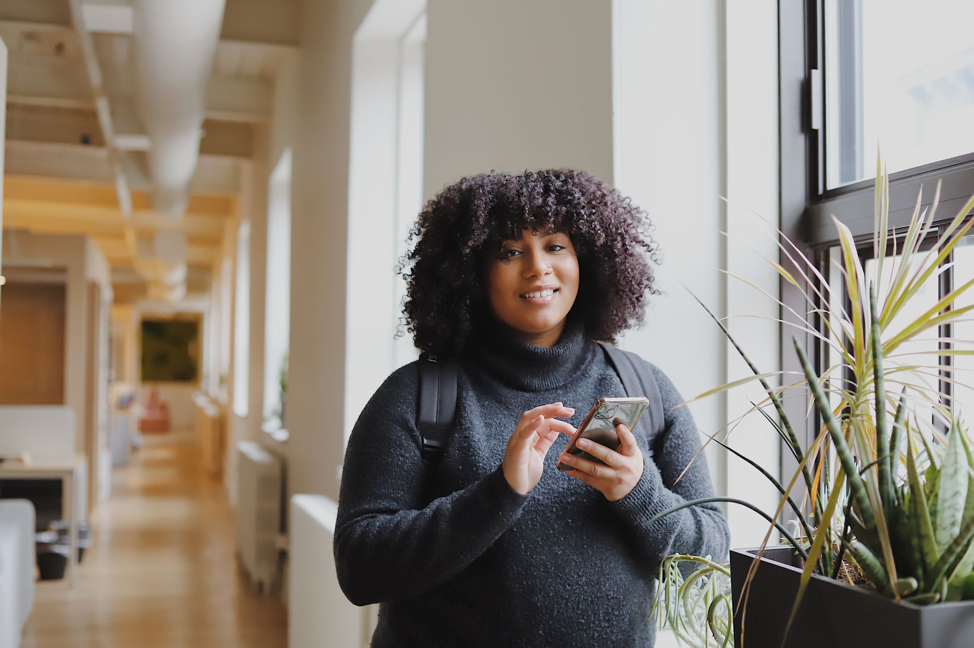 A Black woman with natural curly hair smiles at the camera while typing on her phone in a brightly lit hallway.